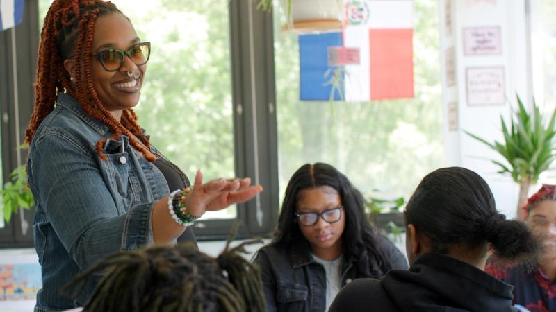 A teacher with orange braids and glasses stands at the front of a bright classroom, gesturing while speaking to a group of seated students.