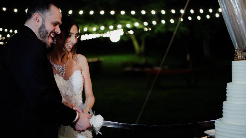 A bride and groom laugh together while cutting their wedding cake at an outdoor evening reception at Springton Manor Farm, with string lights glowing in the background.