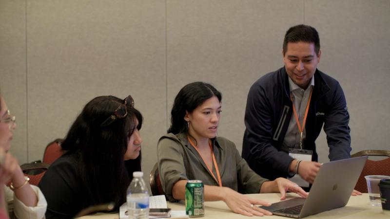 A GivingData conference attendee walks a colleague through something on a laptop at a workshop table, with other participants visible in the background.