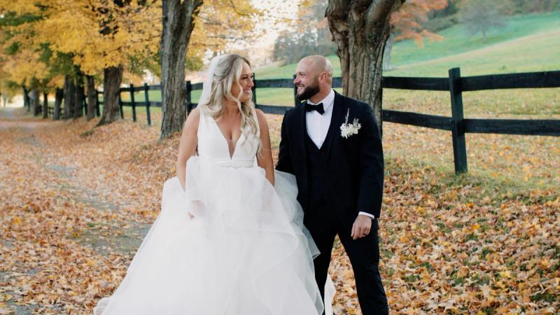 A bride and groom walk hand in hand along a tree-lined path covered in golden autumn leaves, with a black split-rail fence and rolling green hills visible in the background.