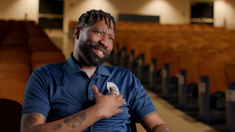A school safety officer in a blue uniform polo speaks during an interview, seated in an auditorium with warm wooden seating visible in the background.