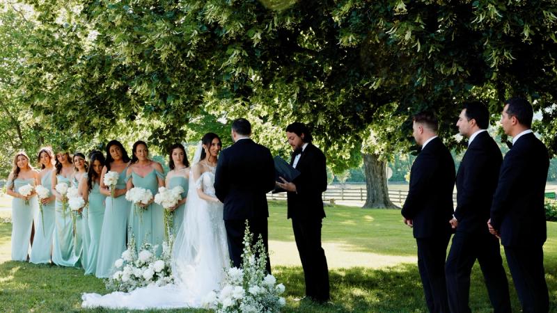 A bride reads her vows from a card while her groom listens with a soft smile during their outdoor ceremony at Springton Manor Farm, surrounded by green trees and summer light.