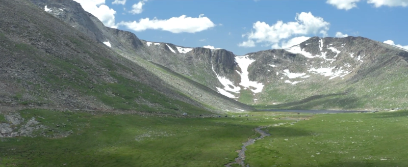 Wide aerial drone shot of a mountain valley in Colorado with a stream running through green alpine meadows, rocky peaks with patches of snow, and a deep blue sky with clouds overhead.