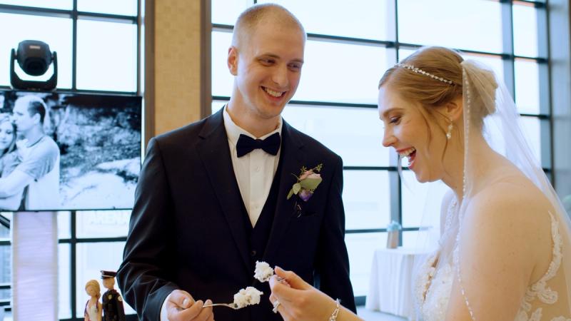 A groom in a black tuxedo and bride in a lace wedding gown laugh together while cutting their wedding cake at Phipps Conservatory, with a large wedding photo displayed on a screen in the background.