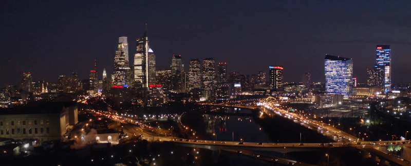 Aerial drone shot of the Philadelphia skyline at night, with the illuminated downtown towers, the Schuylkill River, and lit highway interchanges visible from above.