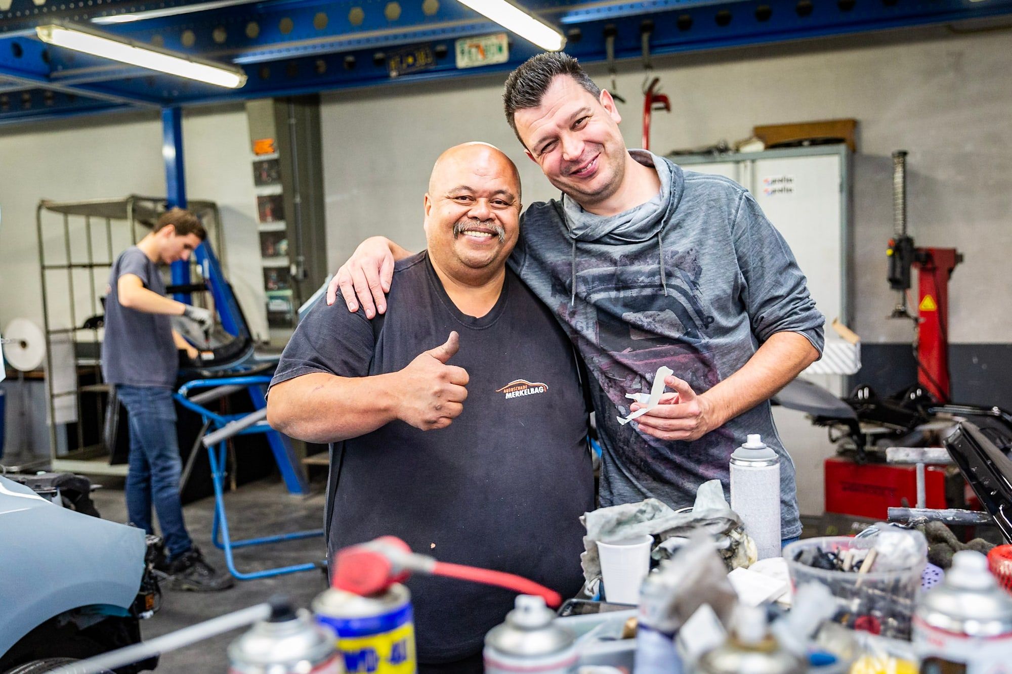 Twee collega's van Autoschade Merkelbag lachend in de werkplaats in Maastricht, met op de achtergrond een collega die aan een auto werkt