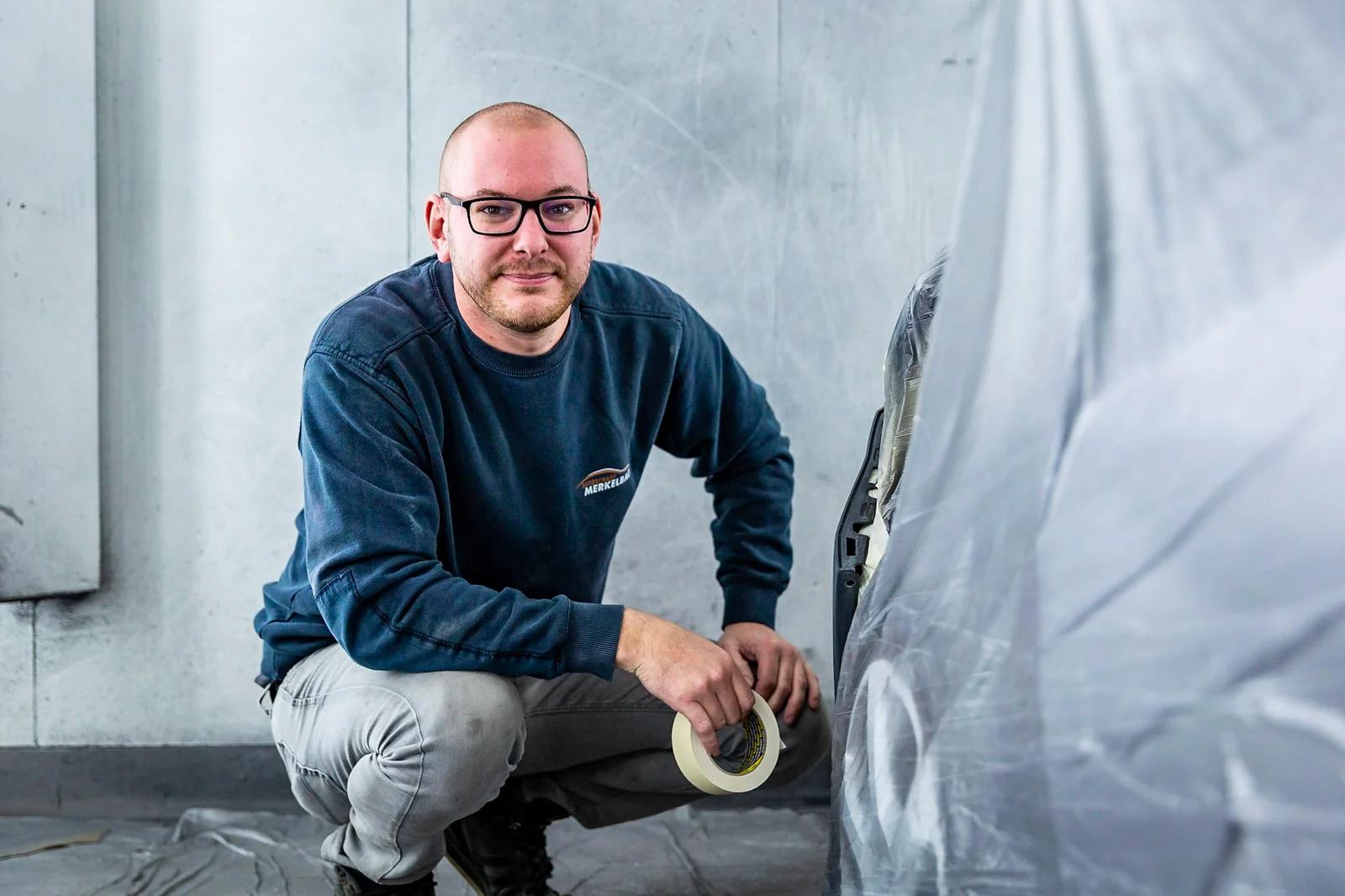 Een spuiter zit gehurkt met een rol afplaktape naast een met folie afgedekte auto in de spuitcabine.