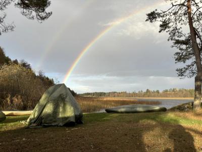Regnbue over et telt og en kano, ved vannet.