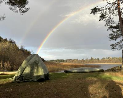 Regnbue over et telt og en kano, ved vannet.