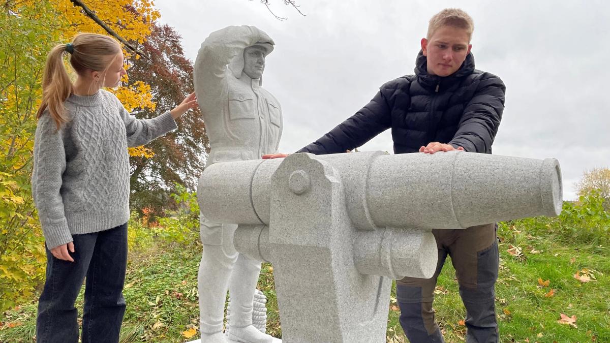 2 elever studerer en kunstinstallasjon, en hvalskytter og en kanon i granitt.