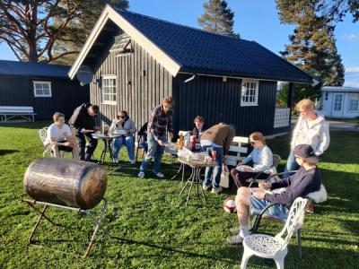 Elever og lærere koser seg med grillmat i kveldssola utenfor en campinghytte.
