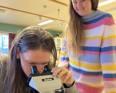 Elever kikker i mikroskop, mens lærer står ved siden og veileder.