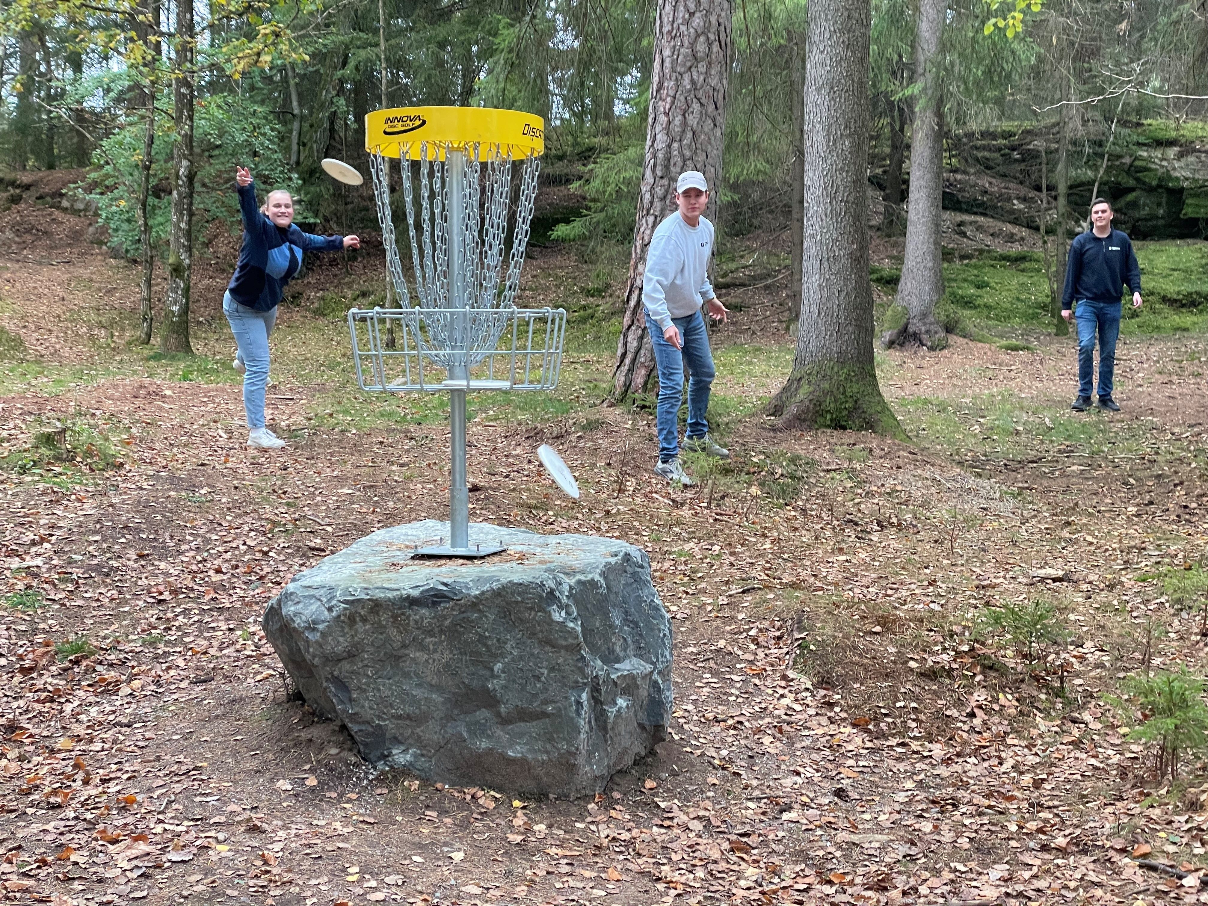 Tre elever forsøker å kaste frisbee i en golfkurv.