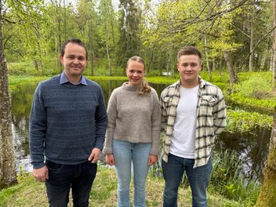 En lærer og to elever oppstilt i en grønn park - foran en dam.