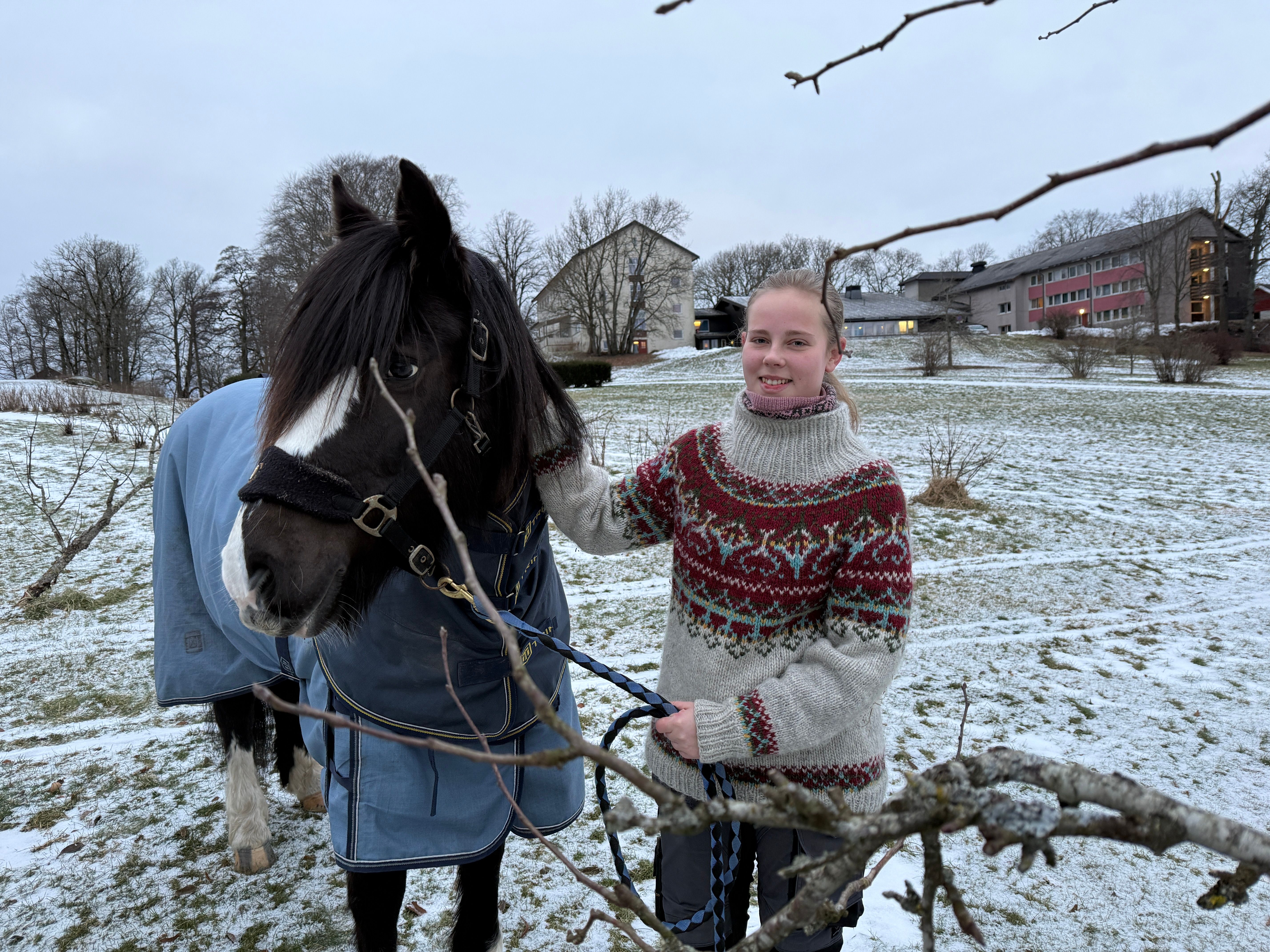 Jente står sammen med hest i en park. Delvis snø på gresset.