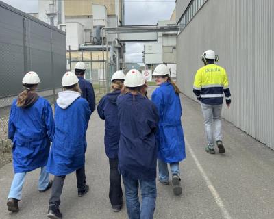 Elever i blå frakker og hvite hjelmer på fabrikk-besøk.