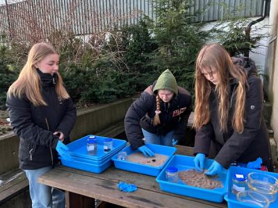 Uteundervisning. Tre elevjenter gjør kjemiforsøk med sand og plastikk i plastkasser.