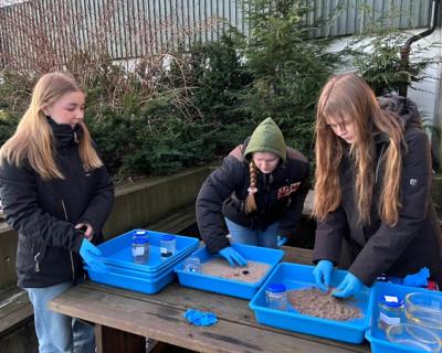 Uteundervisning. Tre elevjenter gjør kjemiforsøk med sand og plastikk i plastkasser.