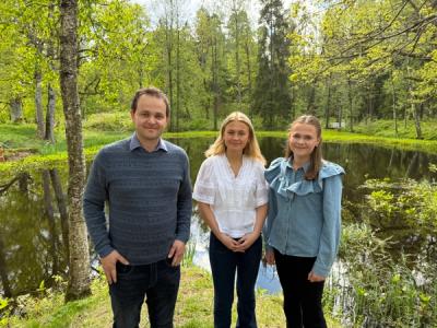 En lærer og to elever oppstilt i en grønn park - foran en dam.