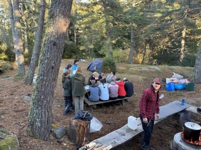 Elever rundt matbordet i skogen. Måltid forberedes på bålet.