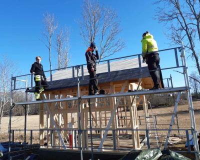 3 elever står på et stilas på et lite bygg og snekrer tak.