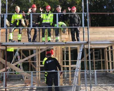 Elever sitter på en stillase og vinker til fotografen.