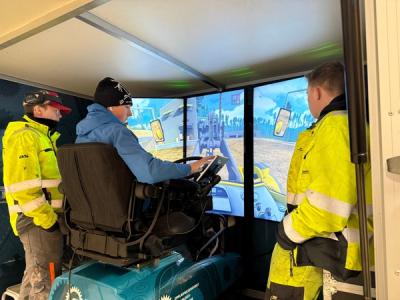 Elev sitter ved en simuleringsmaskin for anleggsmaskiner. Han får veiledning av to elever i gule dresser.