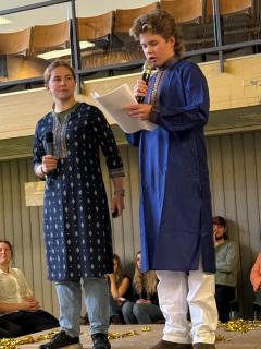 To elever på scene som snakker i mikrofon. De er kledd i sari, en indisk klesdrakt.