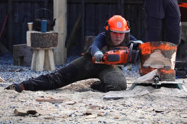 Elev, med hjelm og skogbruksutstyr, bruker motorsag på en vedkubbe.