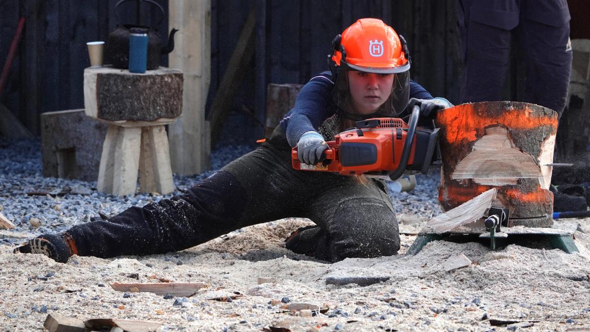 Elev, med hjelm og skogbruksutstyr, bruker motorsag på en vedkubbe.