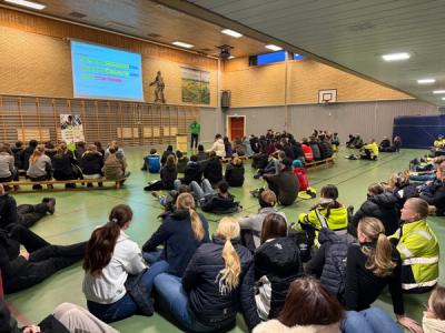 Massevis av elever sitter på gulvet i en gymsal.