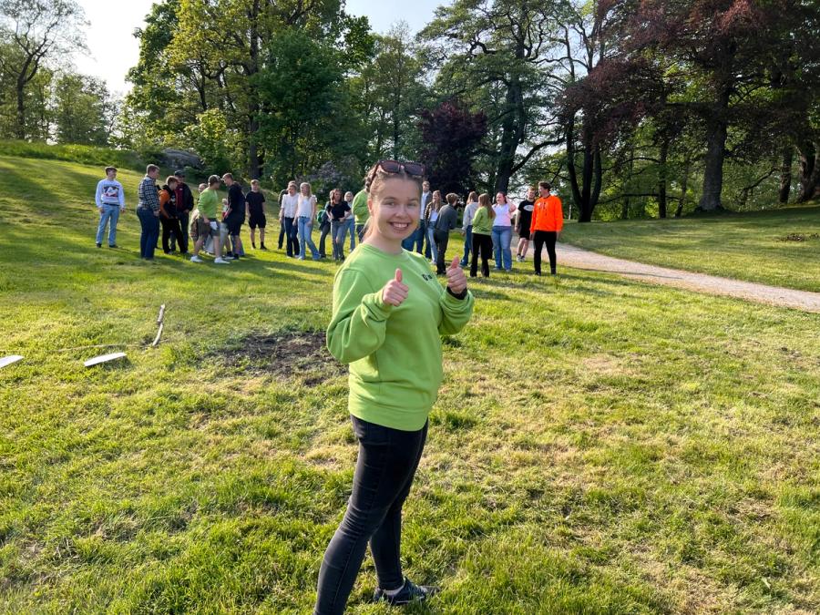 Jente som står med tommel opp i parken, elever i bakgrunnen. 