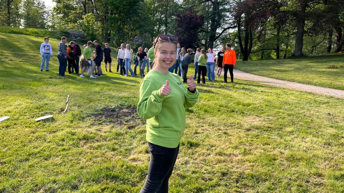 Jente som står med tommel opp i parken, elever i bakgrunnen.