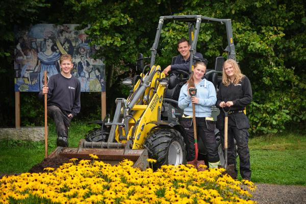 Elever i parken, hjullaster og hageredskaper. Smiler mot camera. 
