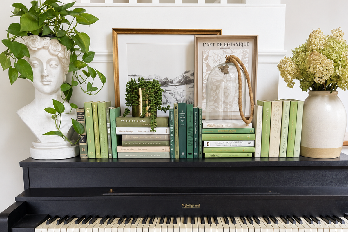 Sage, olive, deep green, and cream books arranged upright and in stacks on a black piano, with leafy plants, framed artwork, and ceramic vases.