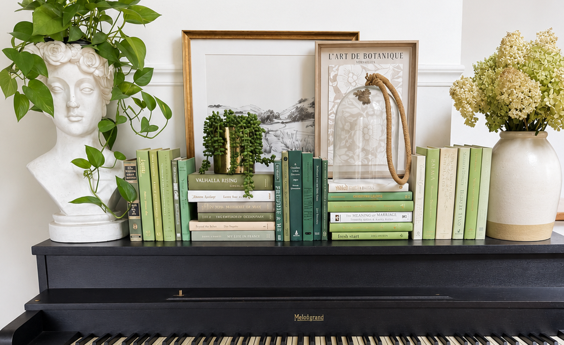 Sage, olive, deep green, and cream books arranged upright and in stacks on a black piano, with leafy plants, framed artwork, and ceramic vases.
