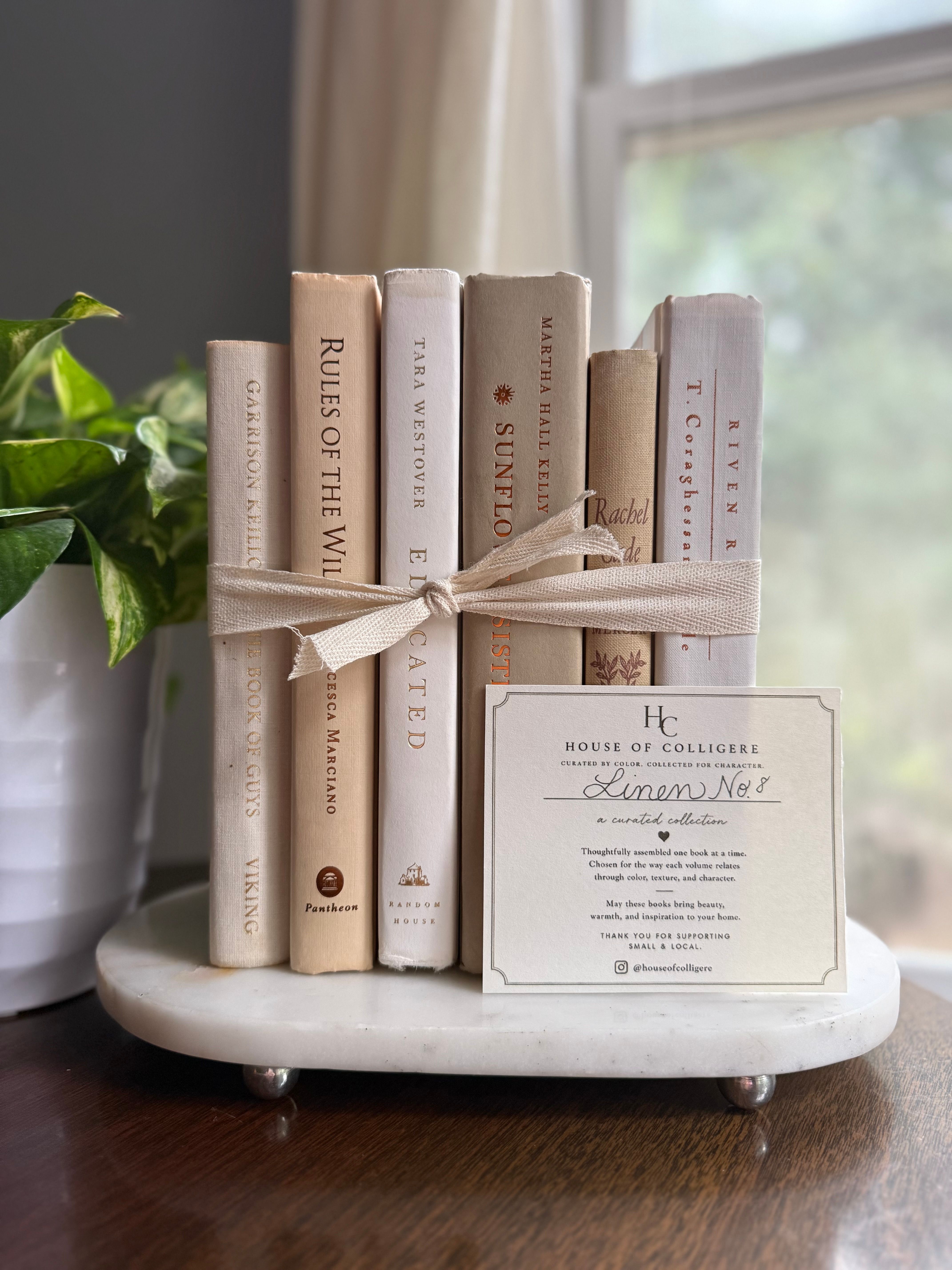 Curated decorative book collection in soft linen-inspired neutrals, featuring ivory, cream, warm beige, and pale taupe clothbound hardcovers with subtle gold, brown, and muted typography.