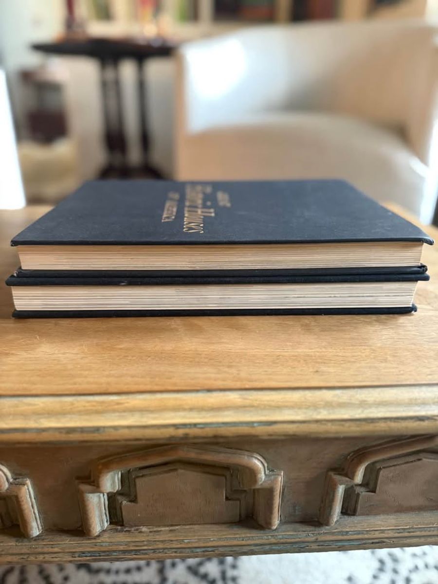 Curated pair of oversized black hardcover books with deep charcoal-black covers, warm aged page edges, and understated gold and cream lettering.