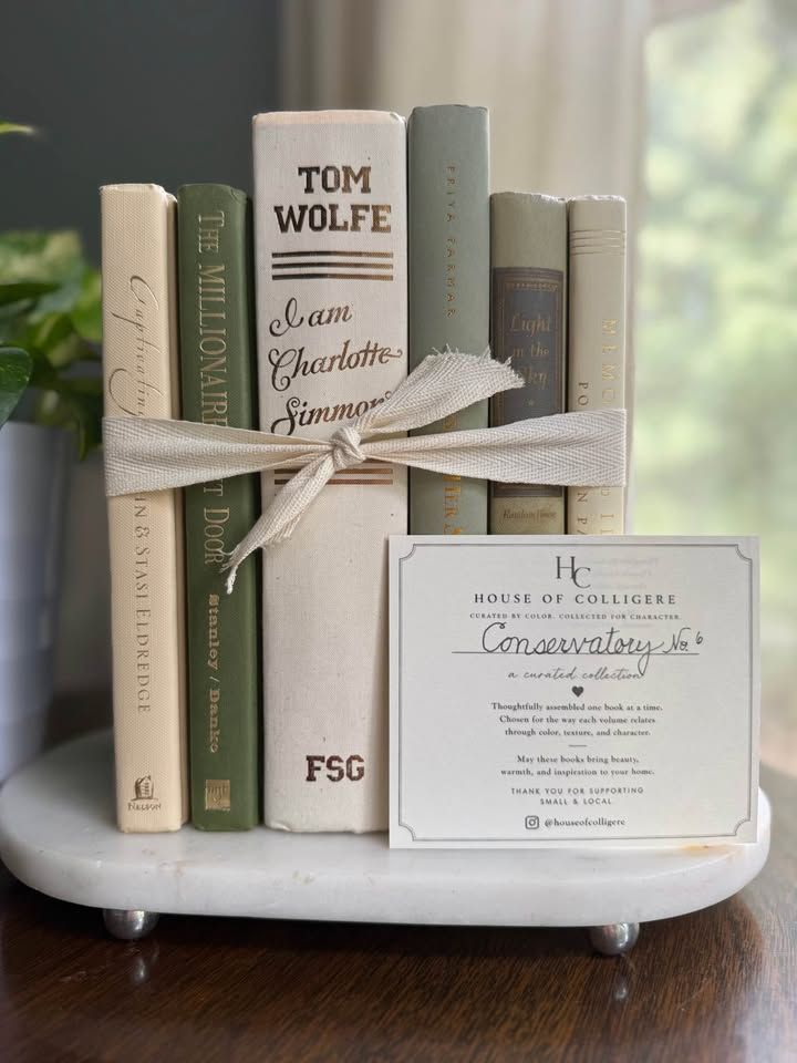 Sage, olive, deep green, and cream books arranged upright and in stacks on a black piano, with leafy plants, framed artwork, and ceramic vases.