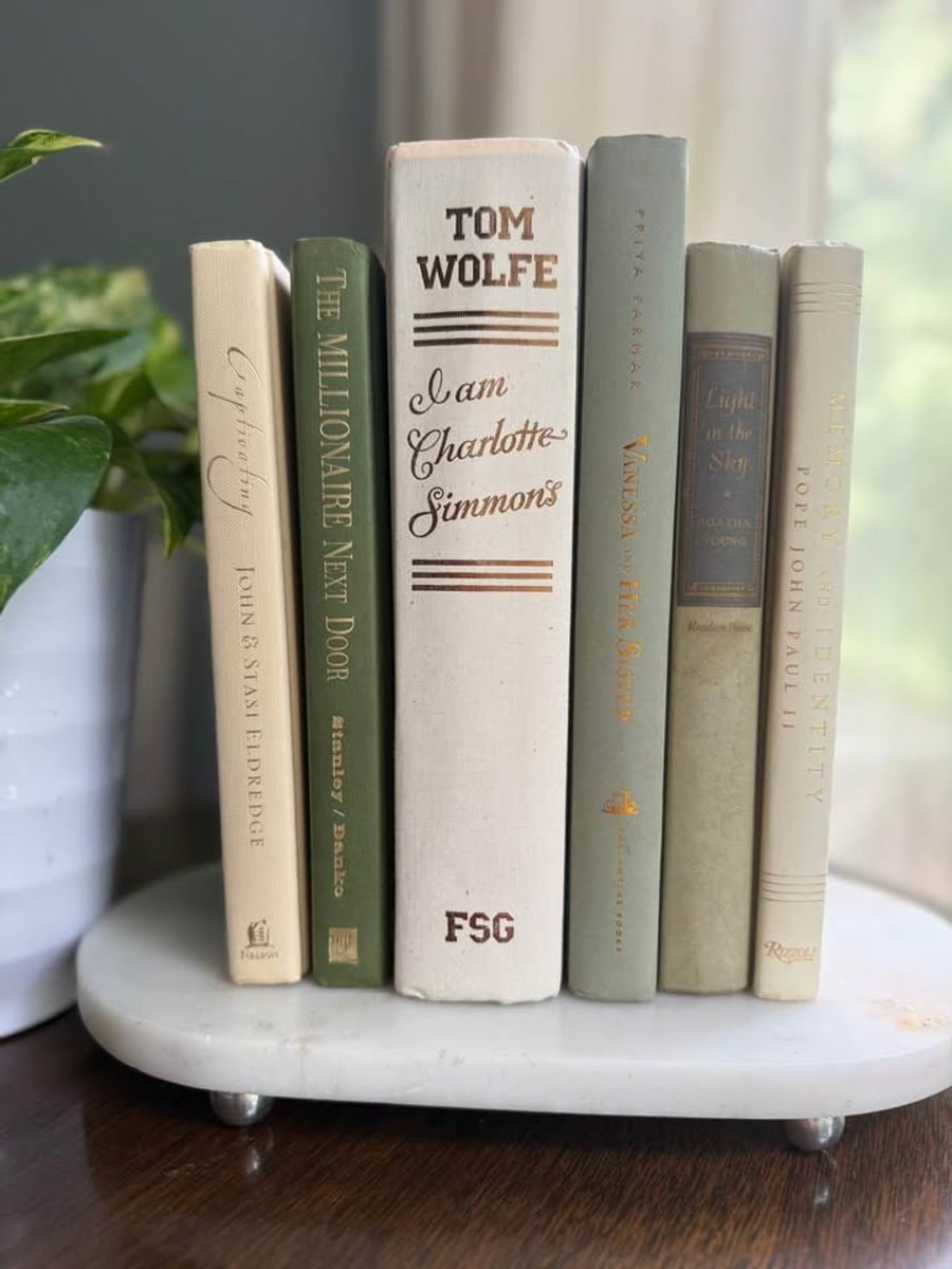 Sage, olive, deep green, and cream books arranged upright and in stacks on a black piano, with leafy plants, framed artwork, and ceramic vases.