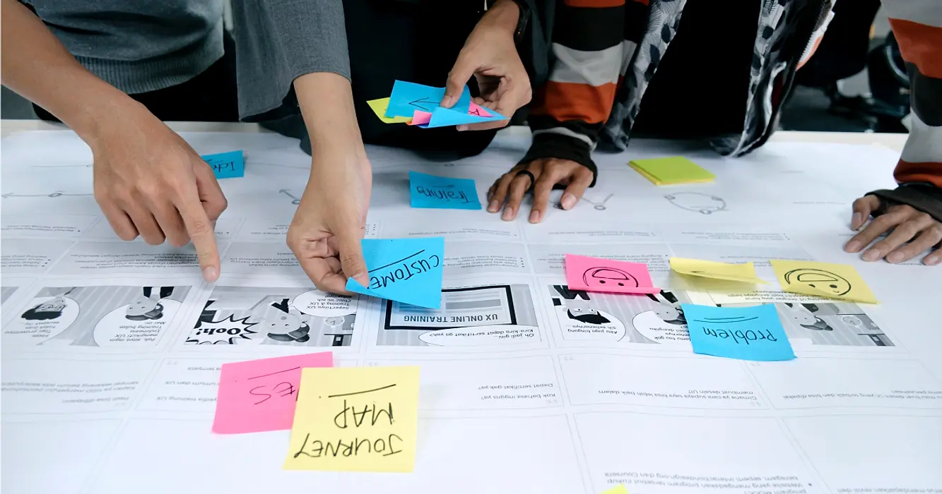 Three people collaborating over table covered in plans and sticky notes planning a digital product.