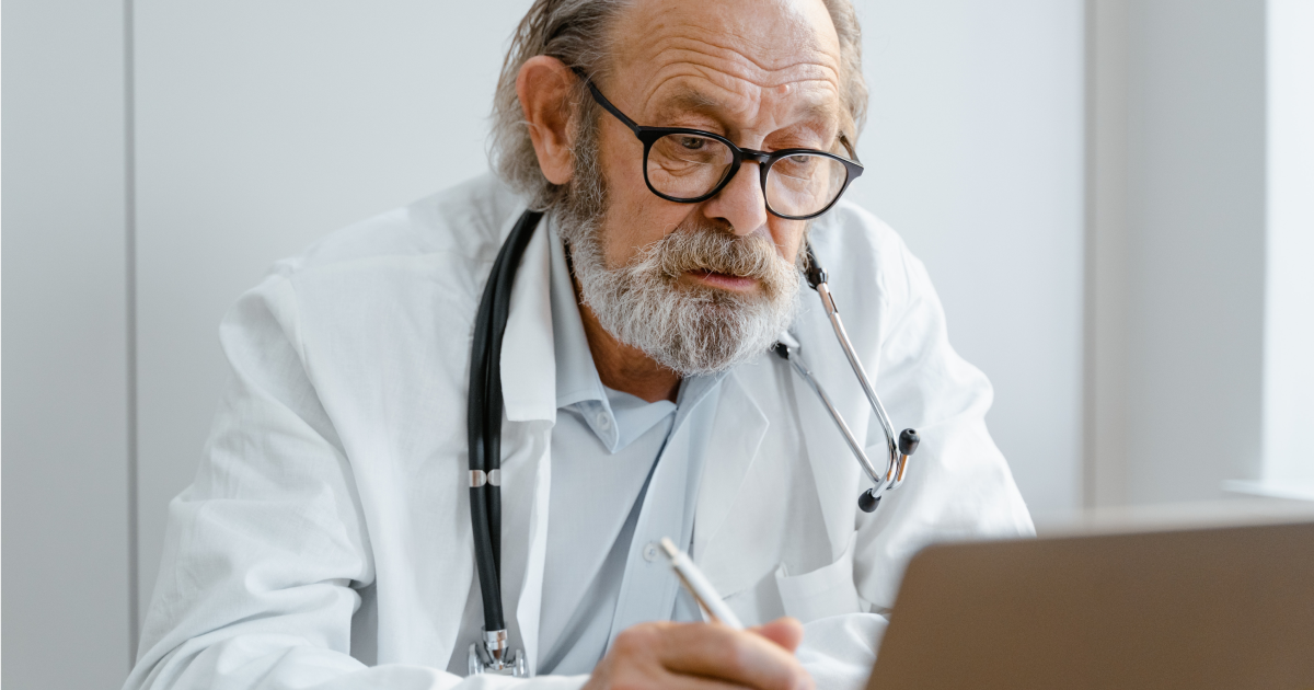 Older male doctor in white lab coat sitting at desk looking at open laptop.