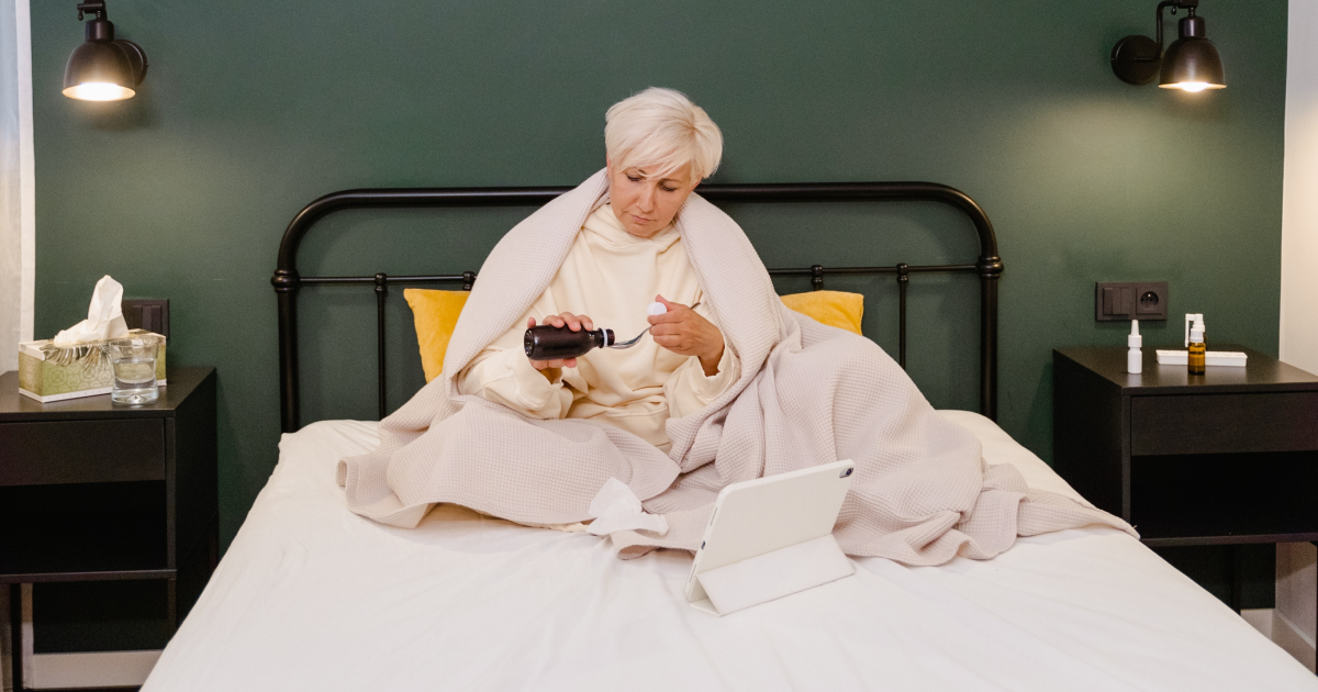 Senior woman sitting in bed pouring medicine into a spoon while having a virtual consultation on tablet device.