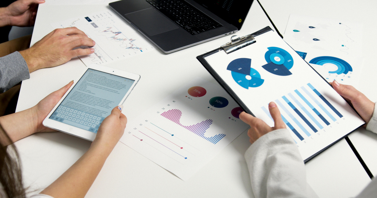 Three people clustered around table looking through charts showing data on printouts and tablet and laptop screens.