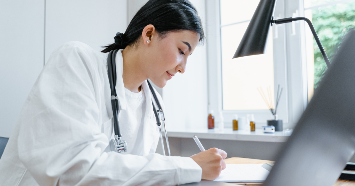 Physician in white lab coat sitting at desk in front of open laptop taking notes on a pad.