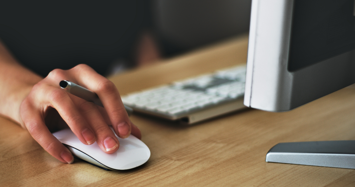 Person's hand using wireless white mouse on wooden table while also holding pen in hand.