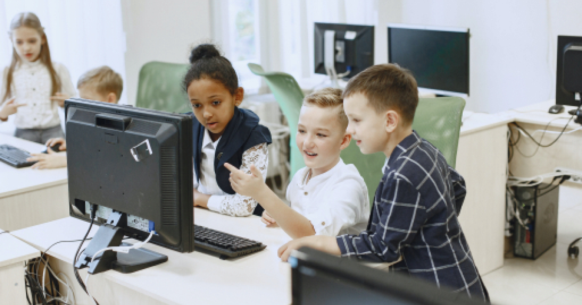 Three kids in computer lab clustered around desktop machine engaging with content on screen.