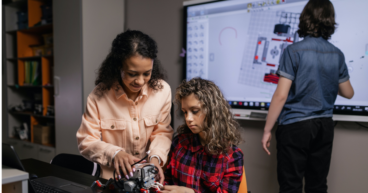 Two middle school aged girls working together to build a robot in a classroom with teacher in background using smartboard.