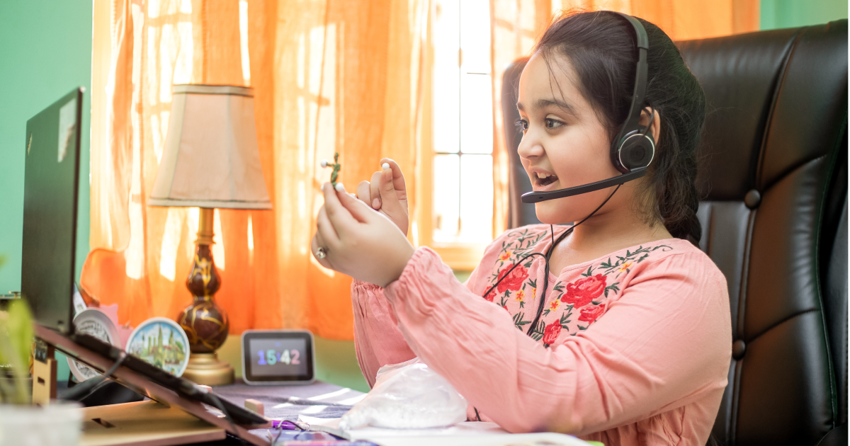 Young girl wearing headset sitting at desk in front of open laptop holding up small figurine to screen while talking.