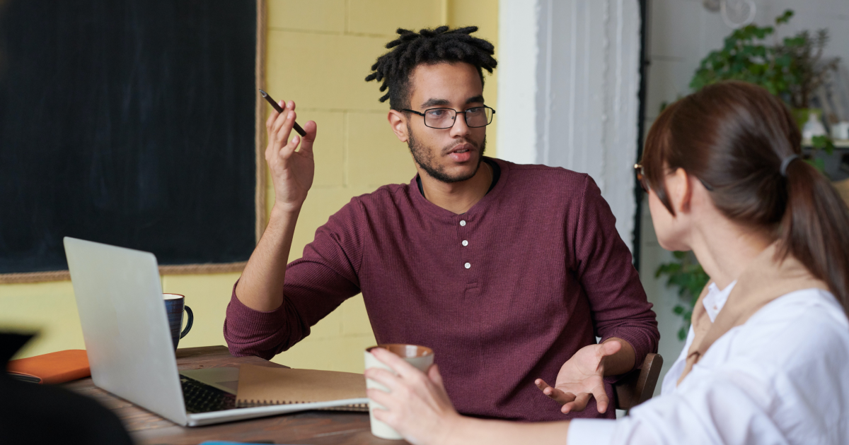 Young man with short dreadlocks and glasses sitting in front of open laptop talking to young woman seated next to him.